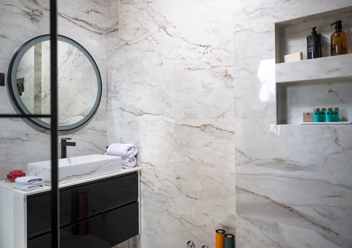A marble-tiled bathroom with a round mirror and walk-in shower